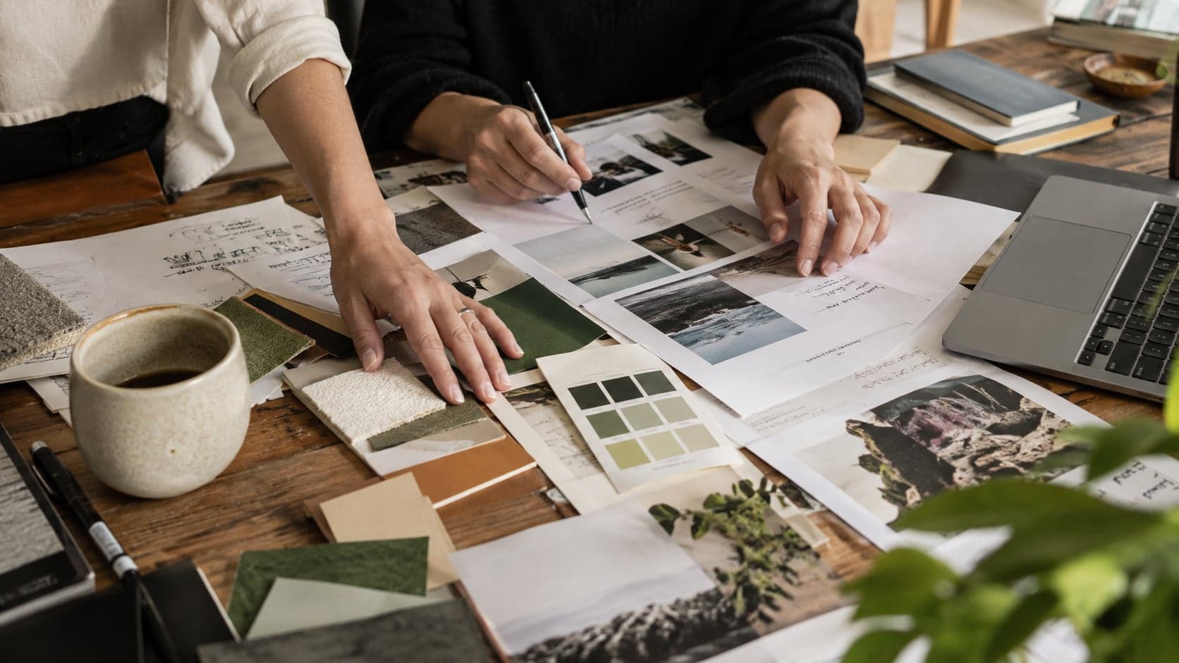 Someone working at a laptop in a bright studio, a sketchbook, printed references and fabric swatches on the table, a wall of pinned images behind